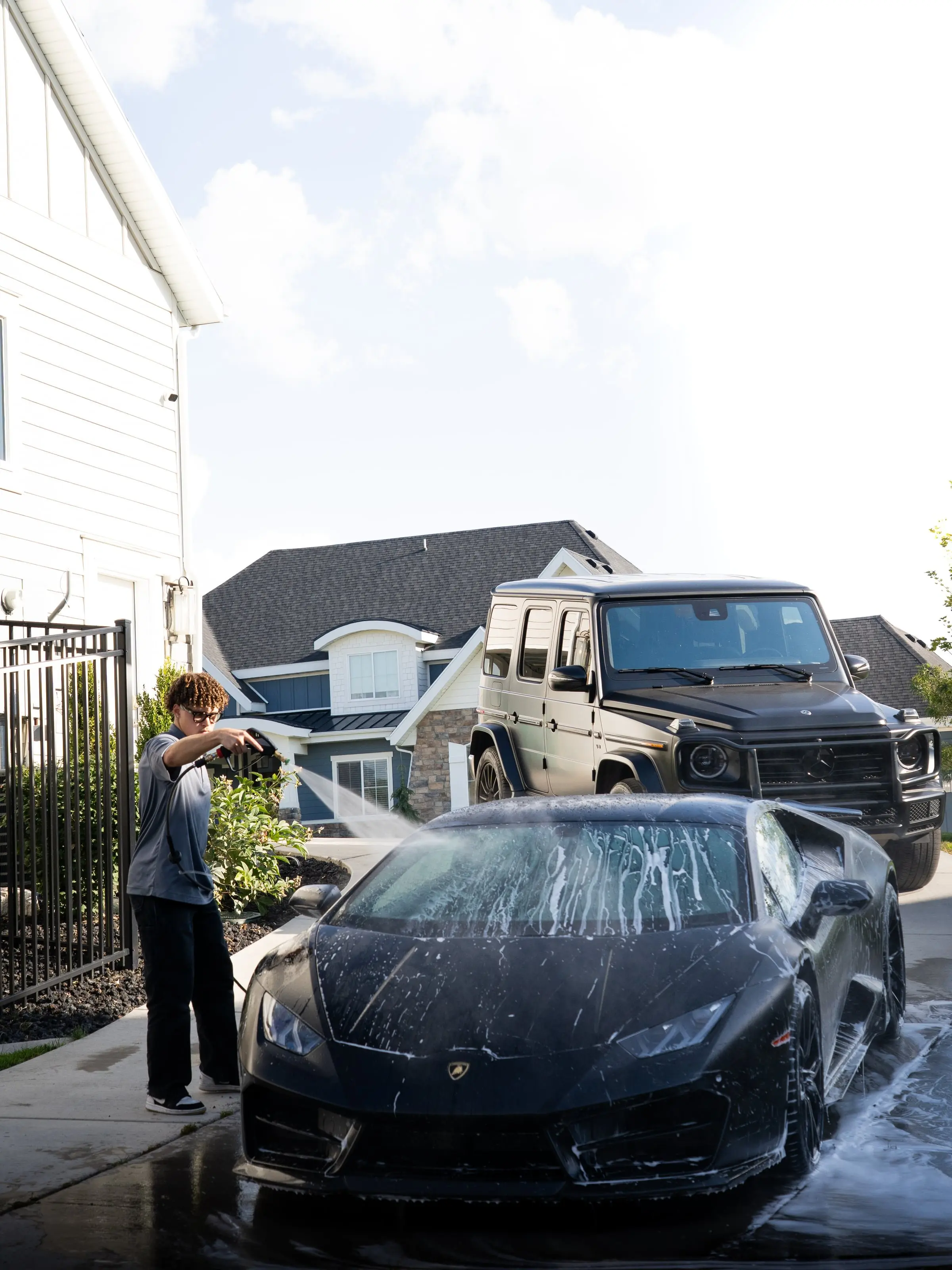 A detailer rinsing a foamed Lamborghini in a client driveway, G-Class behind